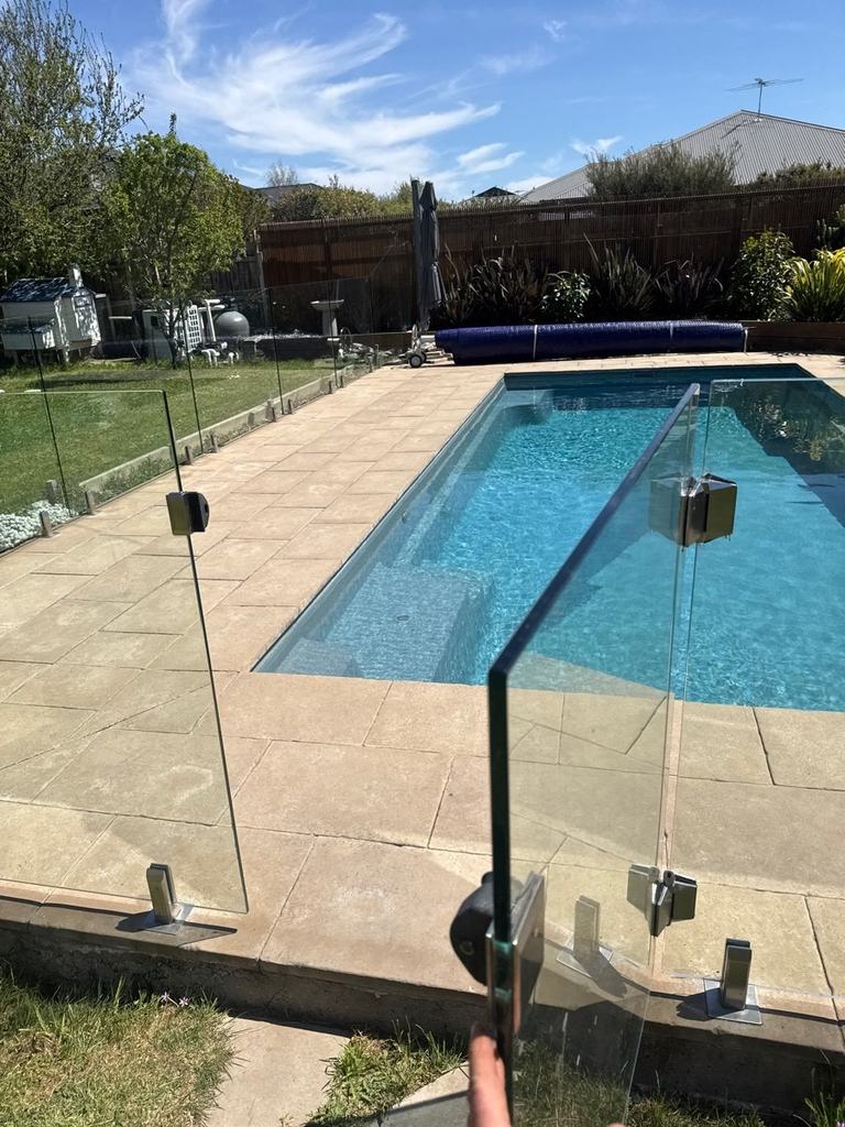 Hand holding a glass pool gate open, with a blue lap pool and paved surround beyond