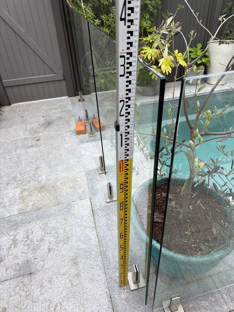 Measuring staff beside a glass pool fence panel with a potted tree close to the barrier