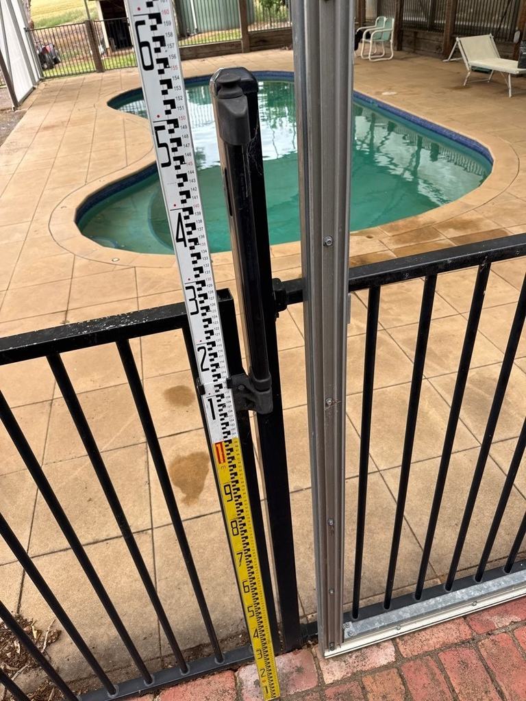 Black tubular pool gate with a metal latch shield and a measuring staff beside the latch
